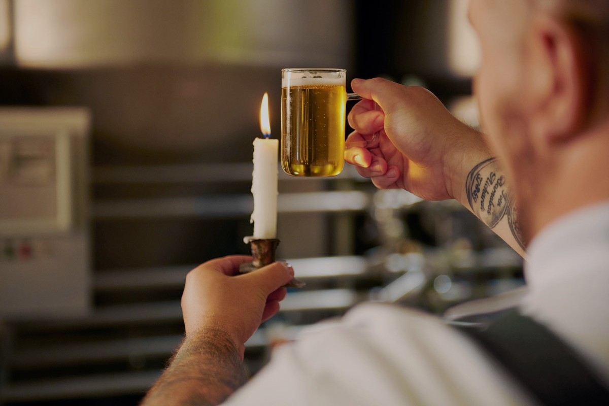 Ein Mann hält ein volles Bierglas nahe an eine brennende Kerze, die er in der anderen Hand hält.