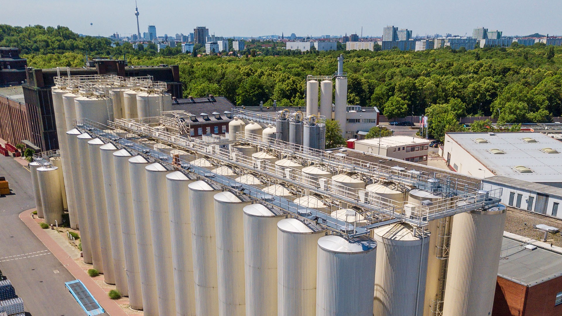 Ein Industriekomplex mit vielen großen Metallsilos und Rohrleitungen, dahinter grüner Wald und Hochhäuser.