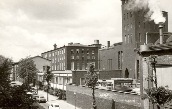 Ein Schwarz-Weiß-Foto zeigt eine industrielle Straßenszene mit Fabrikgebäuden und Autos.