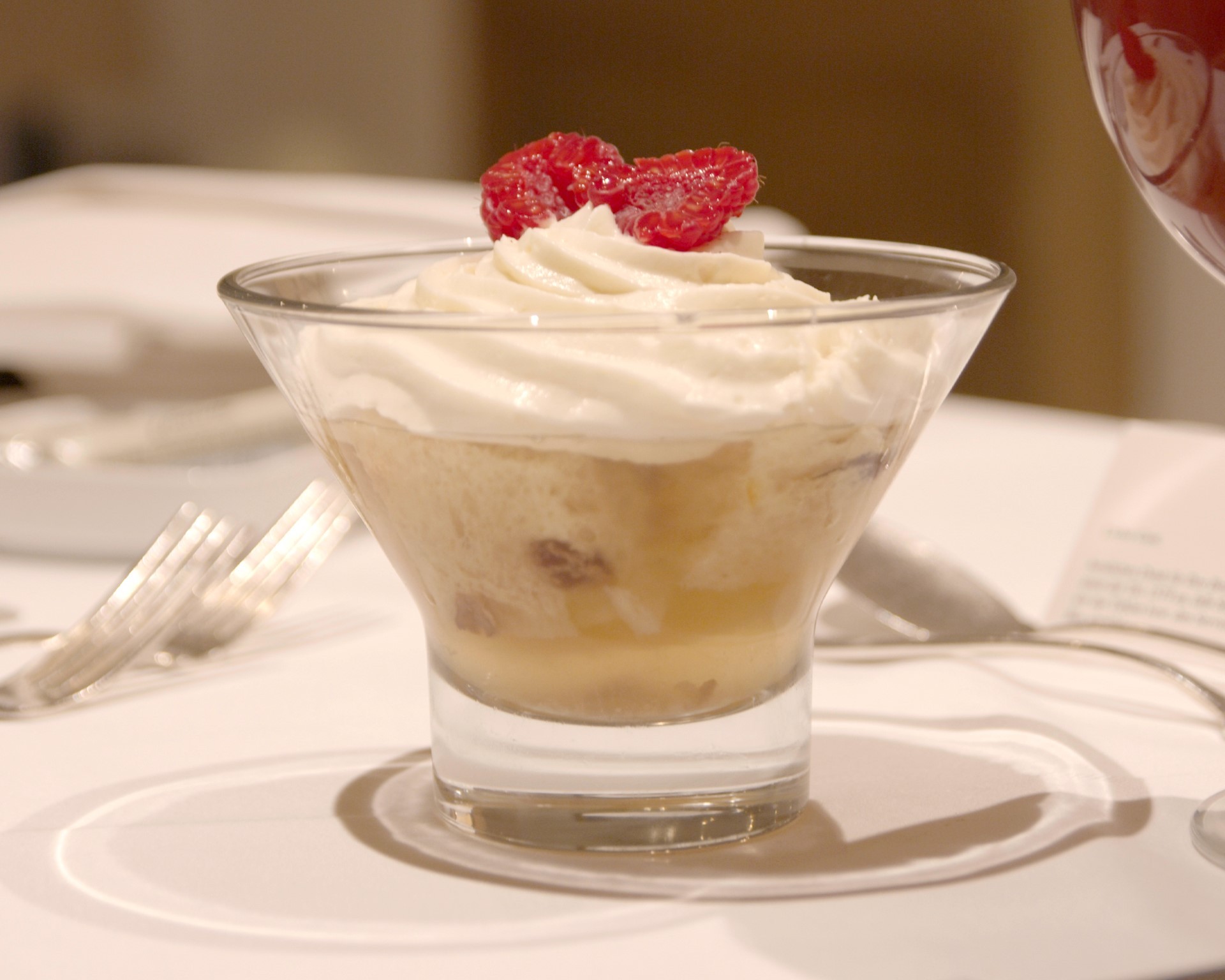 Ein Dessert im Glas steht auf einem Tisch, garniert mit Schlagsahne und Himbeeren oben drauf.