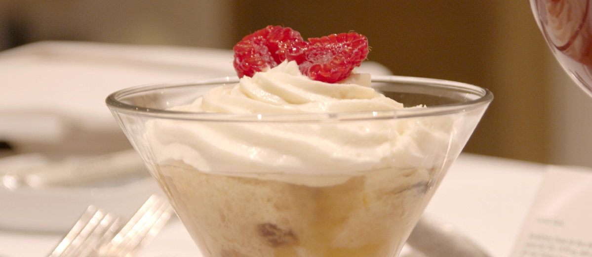 Ein Dessert im Glas steht auf einem Tisch, garniert mit Schlagsahne und Himbeeren oben drauf.
