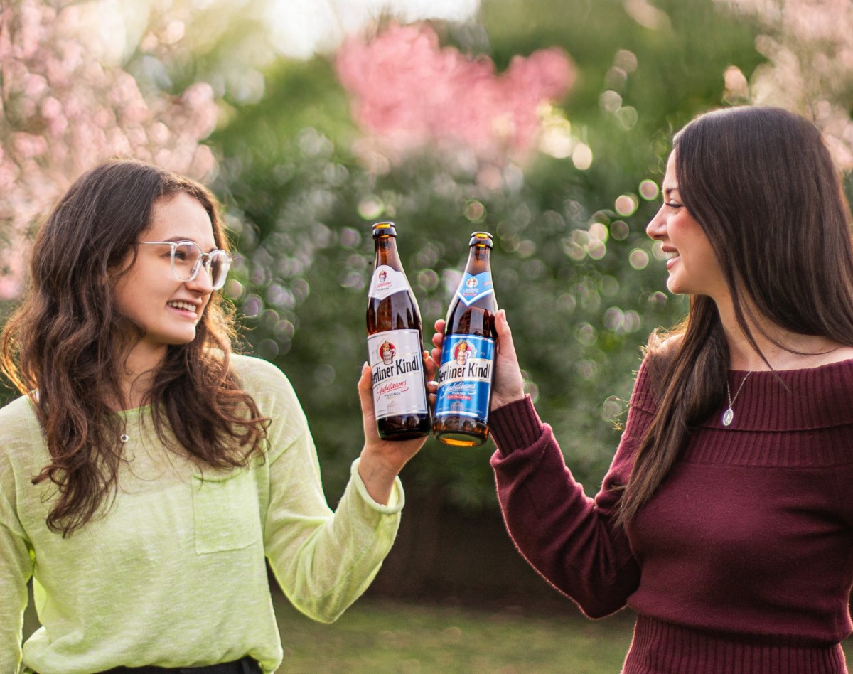Zwei junge Frauen stoßen bei gutem Wetter mit Berliner Kindl Jubiliäums Pilsener und Jubiläums Pilsener Alkoholfrei an.