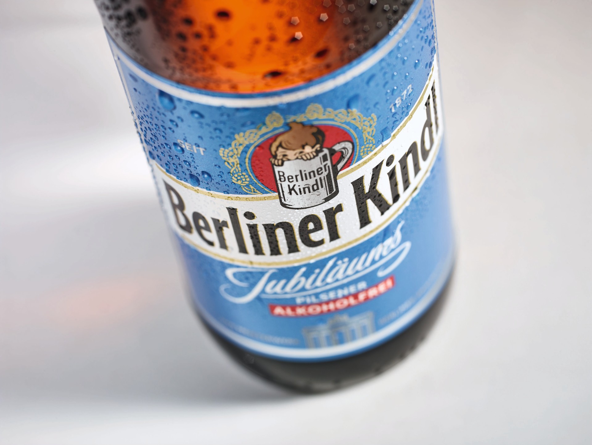 Eine betaute Flasche mit blauem Etikett mit der Aufschrift Berliner Kindl Jubiläums Pilsener Akloholfrei steht auf weißem Untergrund, die Ansicht ist von schräg oben.