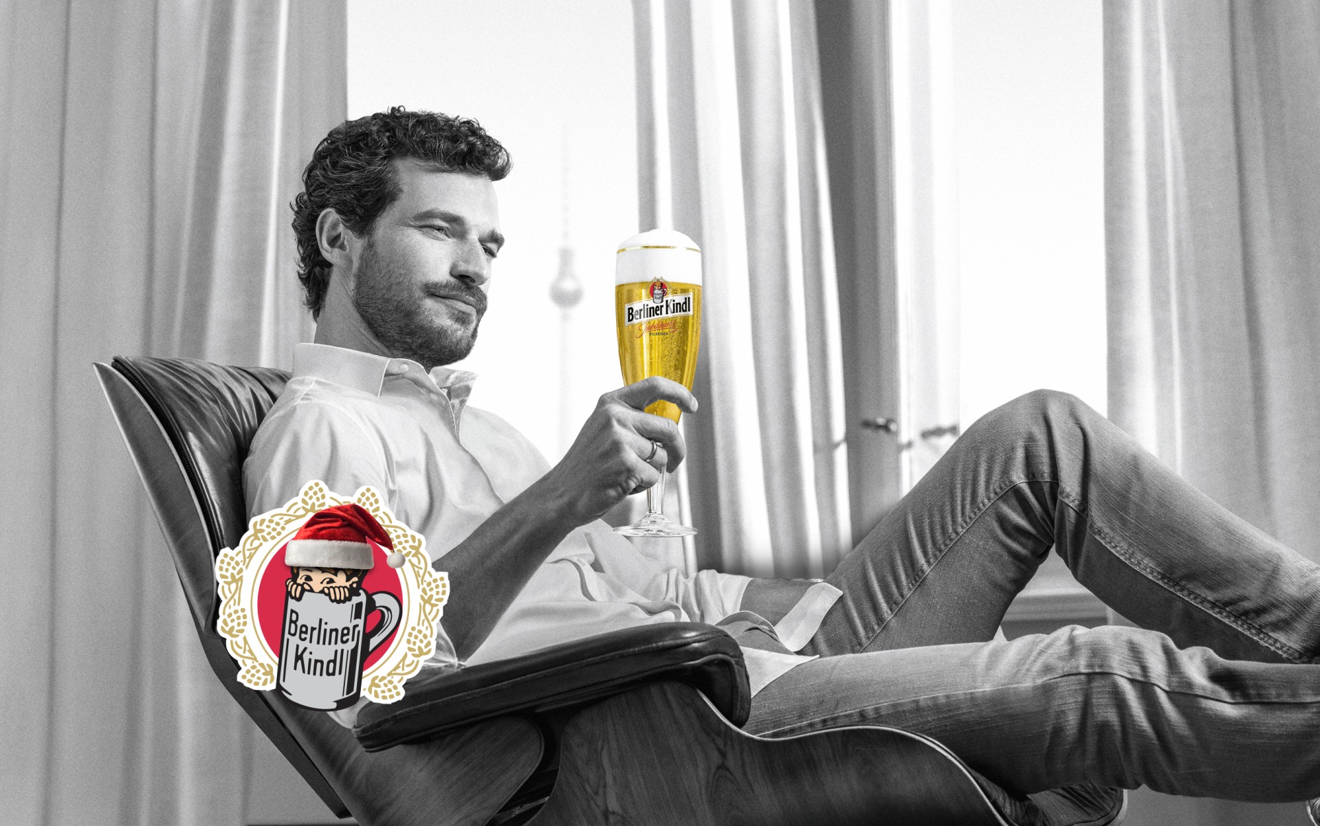 Ein Mann mittleren Alters sitzt lächelnd in einem Sessel und hält ein gefülltes Bierglas. Darunter das Berliner Kindl Logo mit Nikolausmütze.