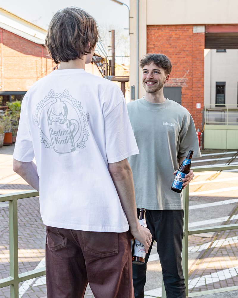 Zwei Männer bekleidet mit Tshirt und Hose stehen auf einem Gebäudevorplatz. Beide halten eine Bierflasche in der Hand.