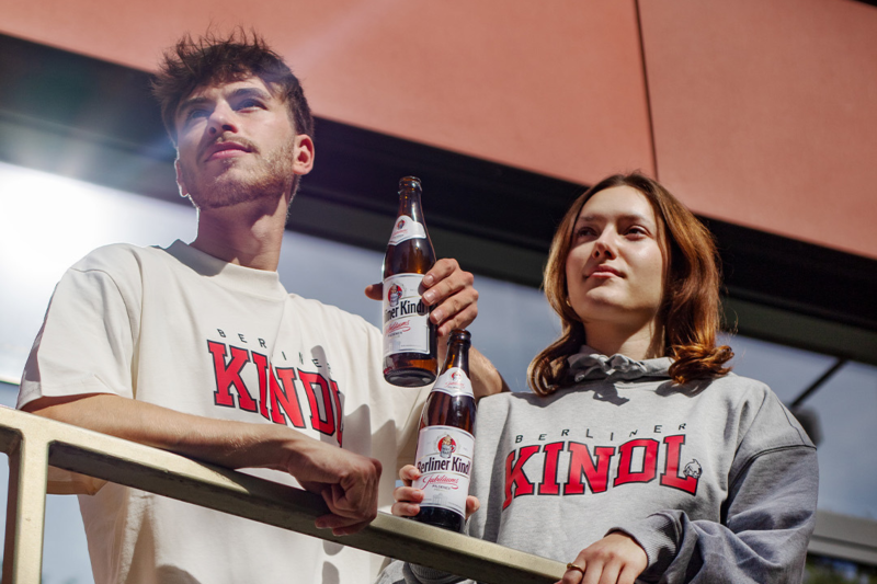Zwei Junge Menschen mit Berliner-Kindl-Sweatshirts halten zwei Bierflaschen im Sonnenlicht.